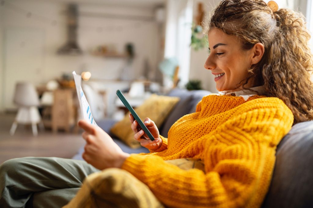 Nasmejana mlada ženska s kodrastimi lasmi, oblečena v udoben rumen pulover, sedi na kavču v svoji dnevni sobi in gleda svoj pametni telefon, medtem ko drži papirnat dokument.