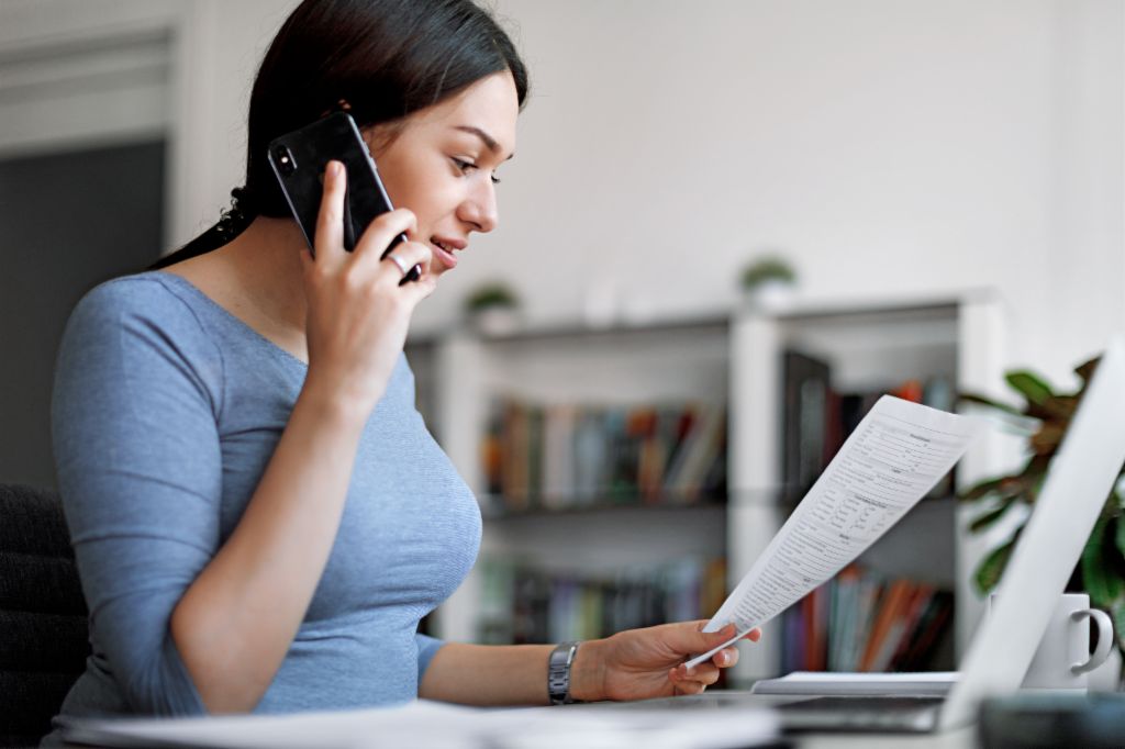 Ženska v modrem topu na mobilnem telefonu, medtem ko drži račun v rokah.