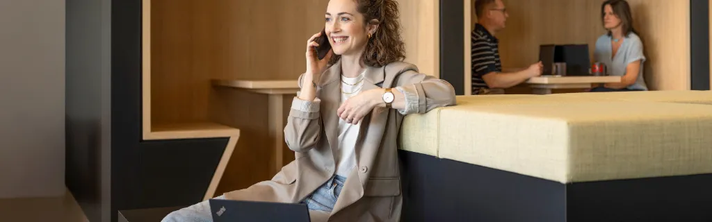Nasmejana oseba sedi v sodobnem pisarniškem salonu, se pogovarja po telefonu in dela na prenosnem računalniku. V ozadju delata še dve osebi.
