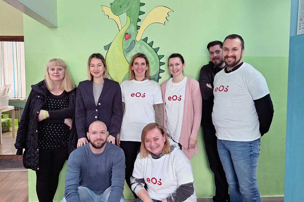 Osem ljudi stoji pred zidom, na katerem je narisan zelen zmaj. Ljudje so postavljeni v dve vrsti: dva spredaj čepita, šest jih stoji zadaj. Štiri osebe nosijo bele majice z rdečim logotipom EOS. Skupina se smehlja v fotoaparat. Na tleh spredaj na desni strani je nekaj steklenic in kartonov z napitki.