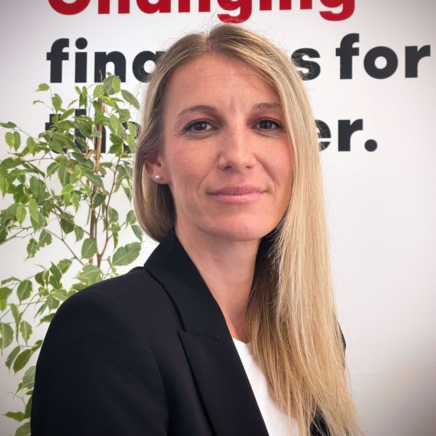 Mirela Lidjan, samostojna pravnica, stoji pred steno z napisom "Changing finances for the better", oblečena v črn suknjič in belo majico, z rastlino v ozadju.