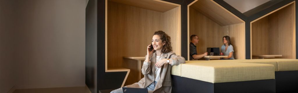 Nasmejana oseba sedi v sodobnem pisarniškem salonu, se pogovarja po telefonu in dela na prenosnem računalniku. V ozadju delata še dve osebi.