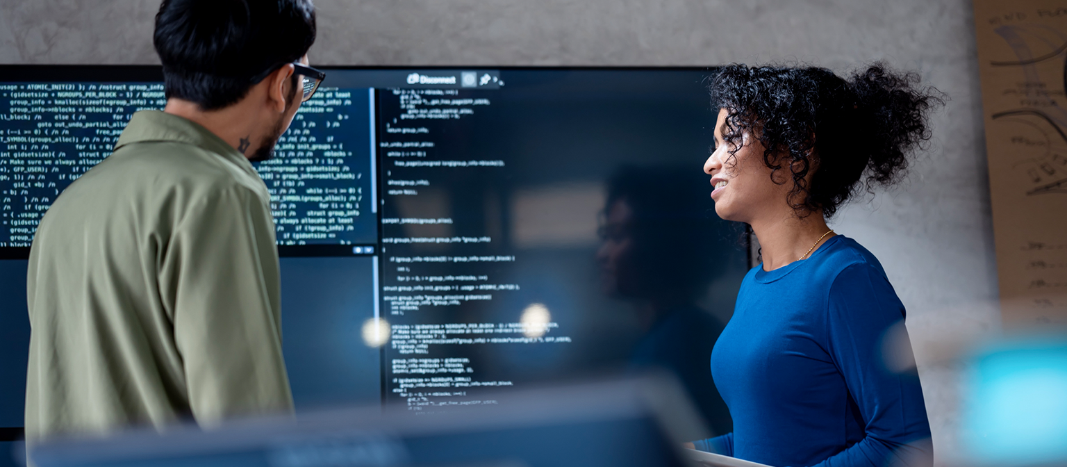 AI v izterjavi dolgov: Na tej sliki dva strokovnjaka skupaj analizirata programski koda za avtomatizirane procese na velikem zaslonu. Scena prikazuje praktično uporabo umetne inteligence (AI) in robotizirane avtomatizacije procesov (RPA) v upravljanju dolgov — primer digitalne transformacije v upravljanju terjatev.