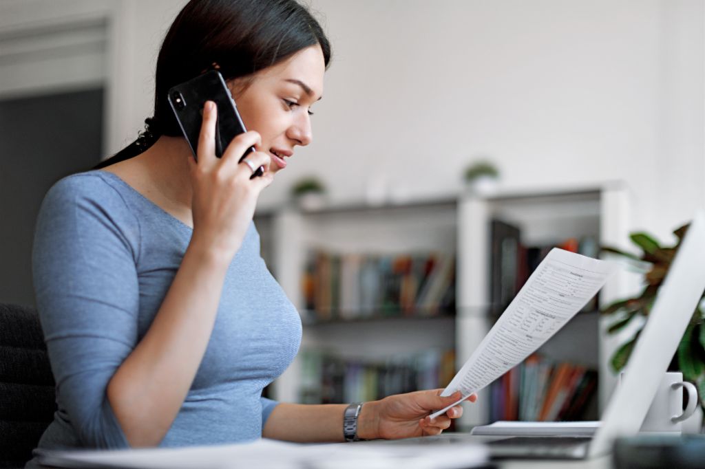 Ženska v modrem topu na mobilnem telefonu, medtem ko drži račun v rokah.